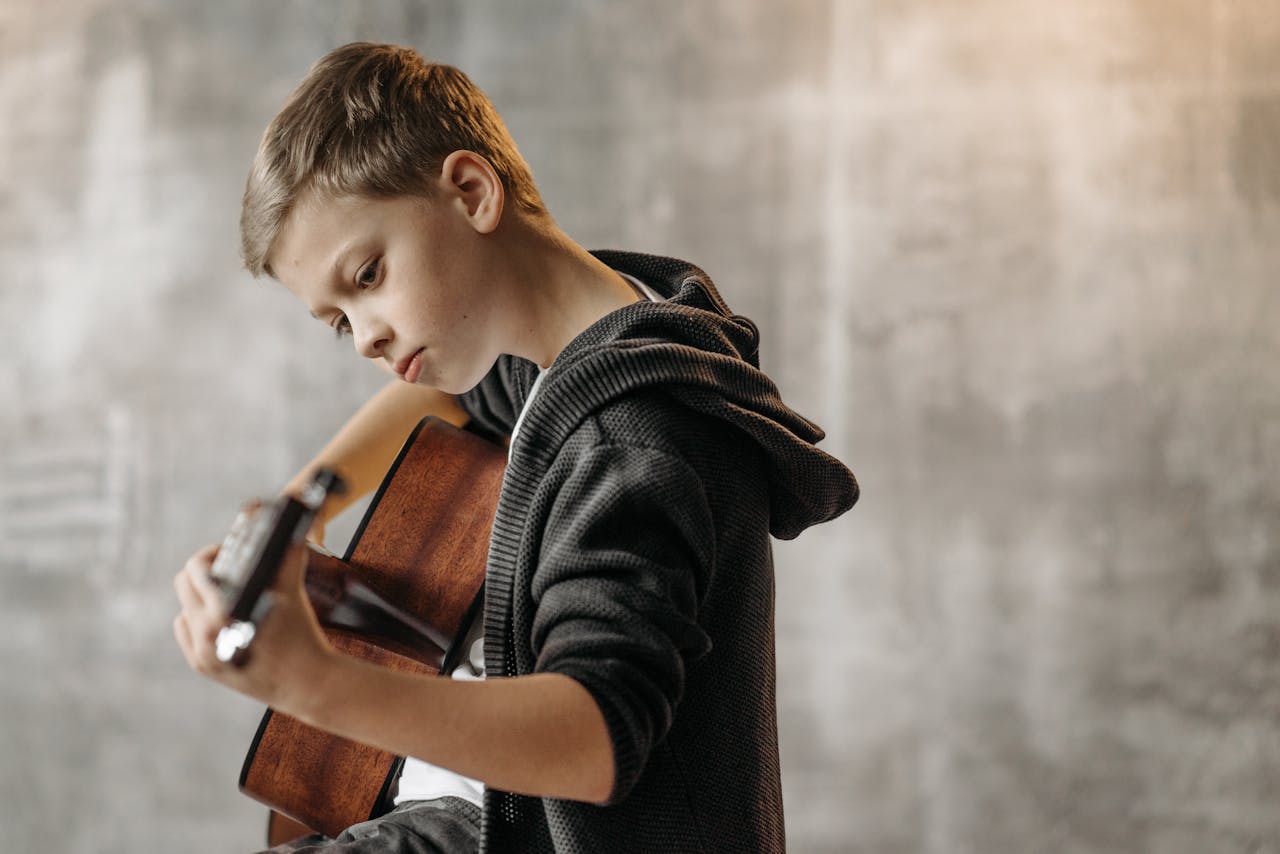A focused young boy playing an acoustic guitar indoors with a neutral background.