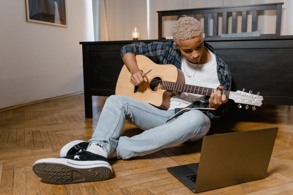 Teen sitting on the floor, playing acoustic guitar beside a laptop, creating music at home.